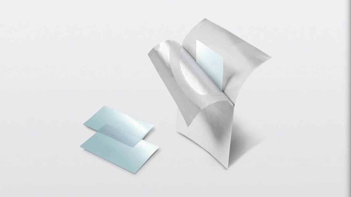 A pair of translucent light blue adhesive patches lies on a light gray surface beside a white, rolled, and partially opened patch that reveals a pale blue inner side.
