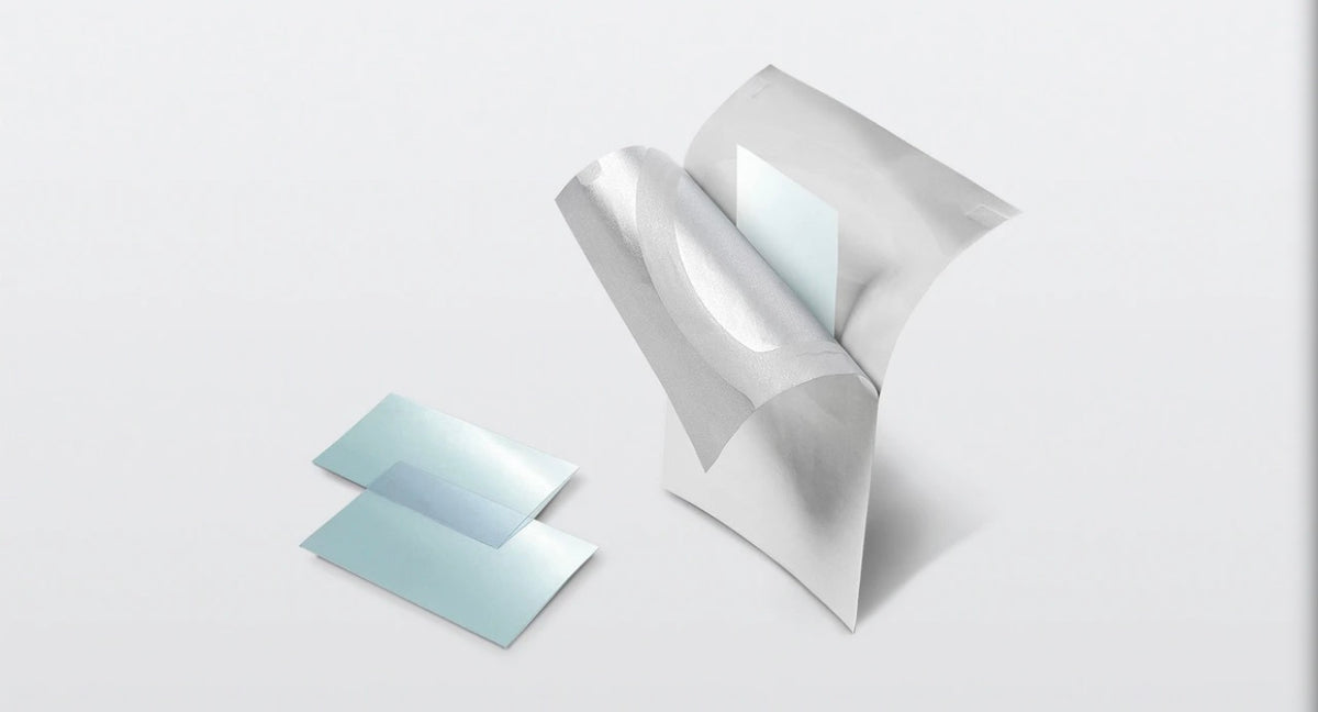 Two translucent light blue square sheets with a metallic silver backing, shown on a clean light gray background. One sheet is standing upright with its backing peeled back, revealing a smooth light blue interior, while the other lies flat beside it.